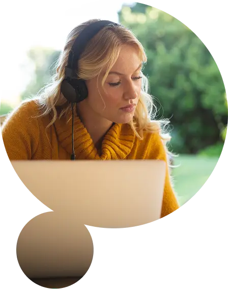 Młoda kobieta w żółtym swetrze, w słuchawkach, uczy się przy laptopie i książce przy biurku przy oknie. Zieleń za oknem tworzy spokojne tło.