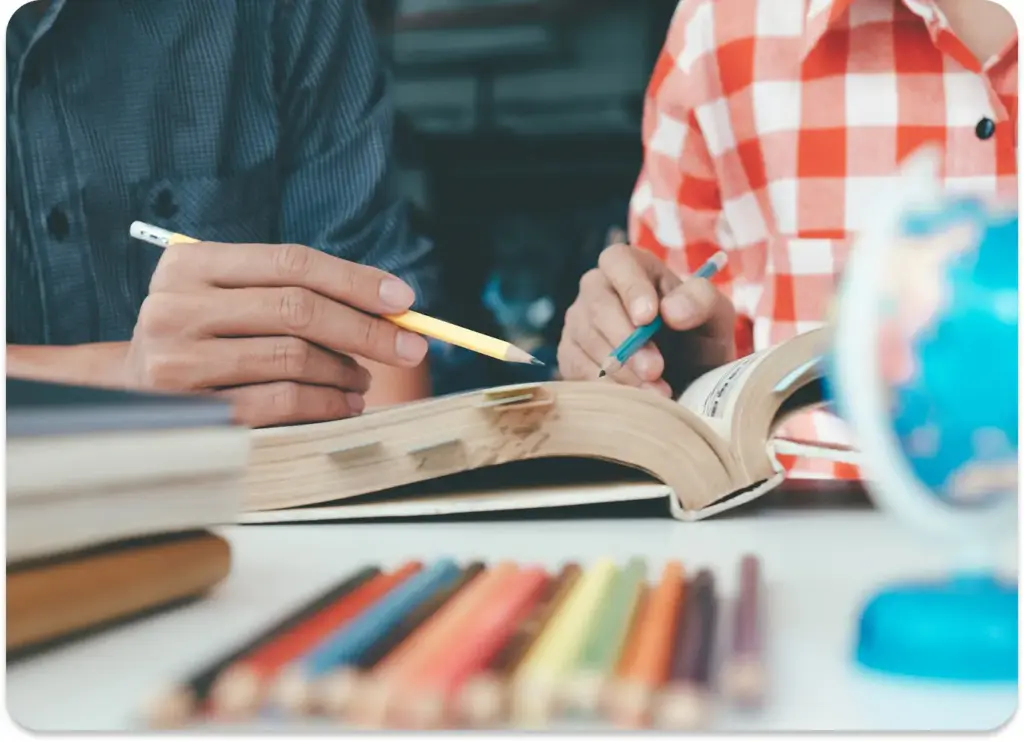 Dwie osoby siedzące przy stole studiują otwartą książkę, trzymając w rękach ołówki. Widoczne są kolorowe ołówki i mały globus, które tworzą atmosferę nauki.