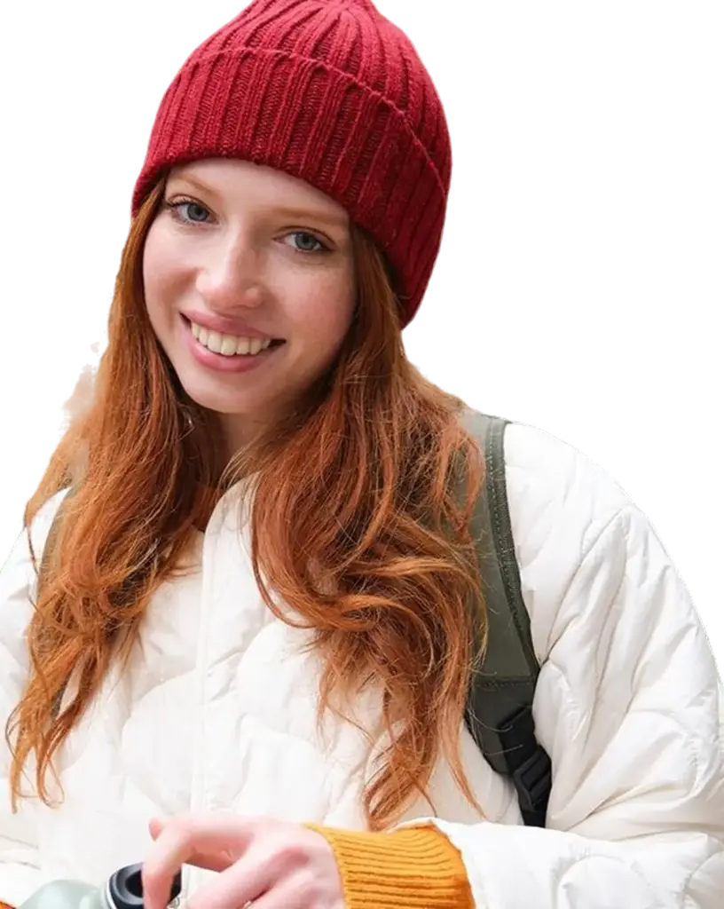 Młoda kobieta o długich rudych włosach uśmiecha się ciepło, mając na sobie czerwoną czapkę z dzianiny i białą kurtkę. Trzyma butelkę wody, emanując radością i spokojem.