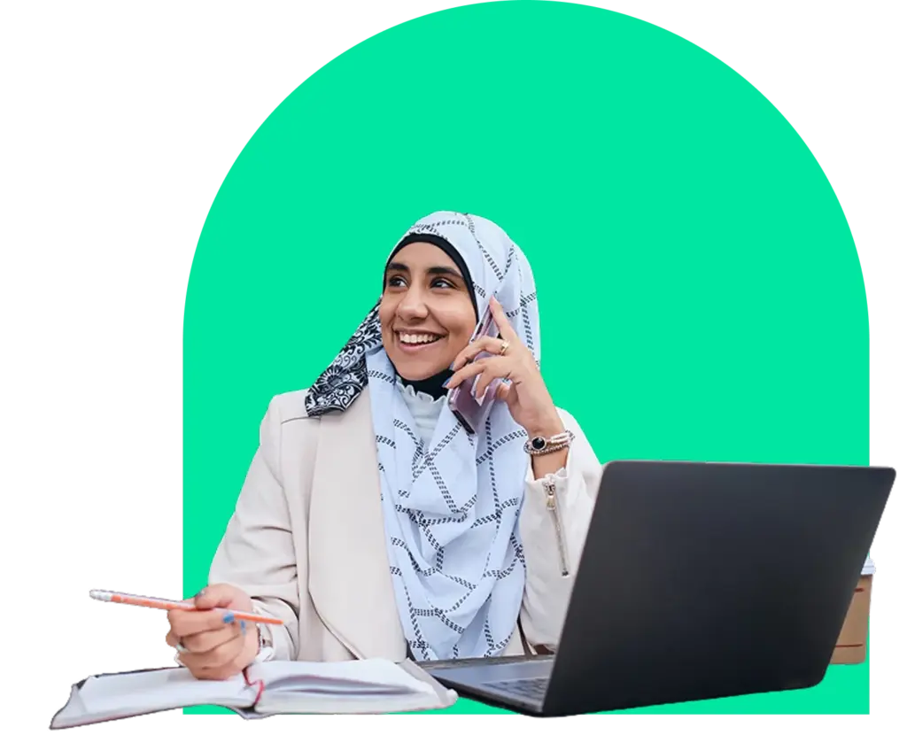 Kobieta w hidżabie i jasnej marynarce uśmiecha się podczas rozmowy telefonicznej. Trzyma długopis, a przed sobą ma otwarty notatnik i laptop, co nadaje jej produktywny wygląd.